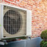 Air conditioning unit on a brick wall with green foliage, from Cardinal Heating, Cooling, Plumbing & Electric.