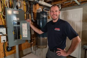 Smiling technician next to an electrical panel and pipes in a basement for Cardinal Heating, Cooling, Plumbing & Electric.