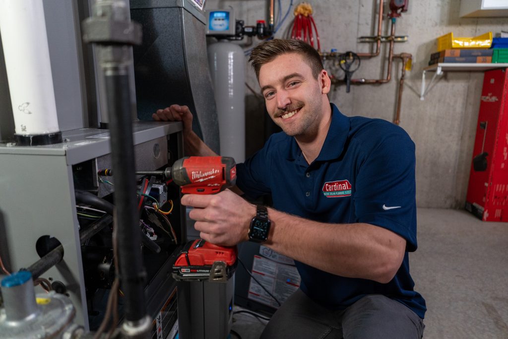 Technician with Cardinal logo using a power tool near HVAC equipment in a professional workshop setting.
