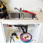 Stainless steel kitchen sink with plumbing tools, including a red plunger and colorful hoses, emphasizing plumbing maintenance and repair services.