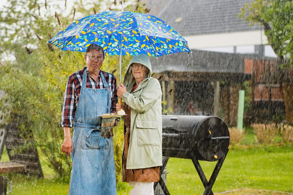 Elderly couple standing under a colorful umbrella in the rain, holding a plate, with a grill in the background, symbolizing summer grilling and outdoor activities during unpredictable weather.