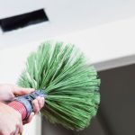 Person using a green duct cleaning brush to clean an air vent, emphasizing air duct maintenance for improved indoor air quality.